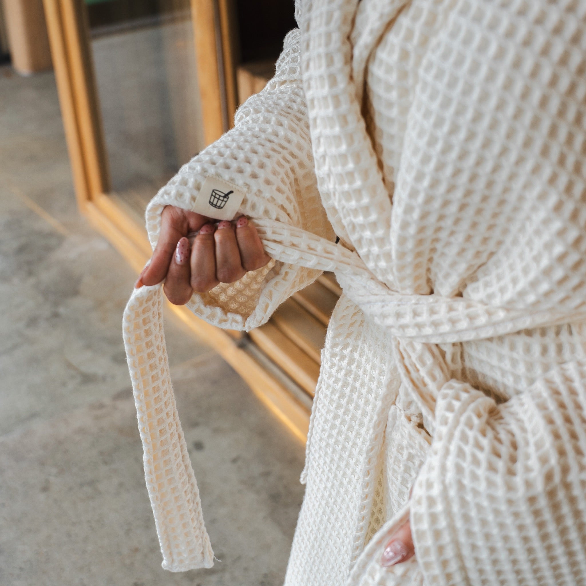 Tying the belt of The Sauna Robe in Arctic Ivory, waffle weave with woven Cult of Sauna cuff label