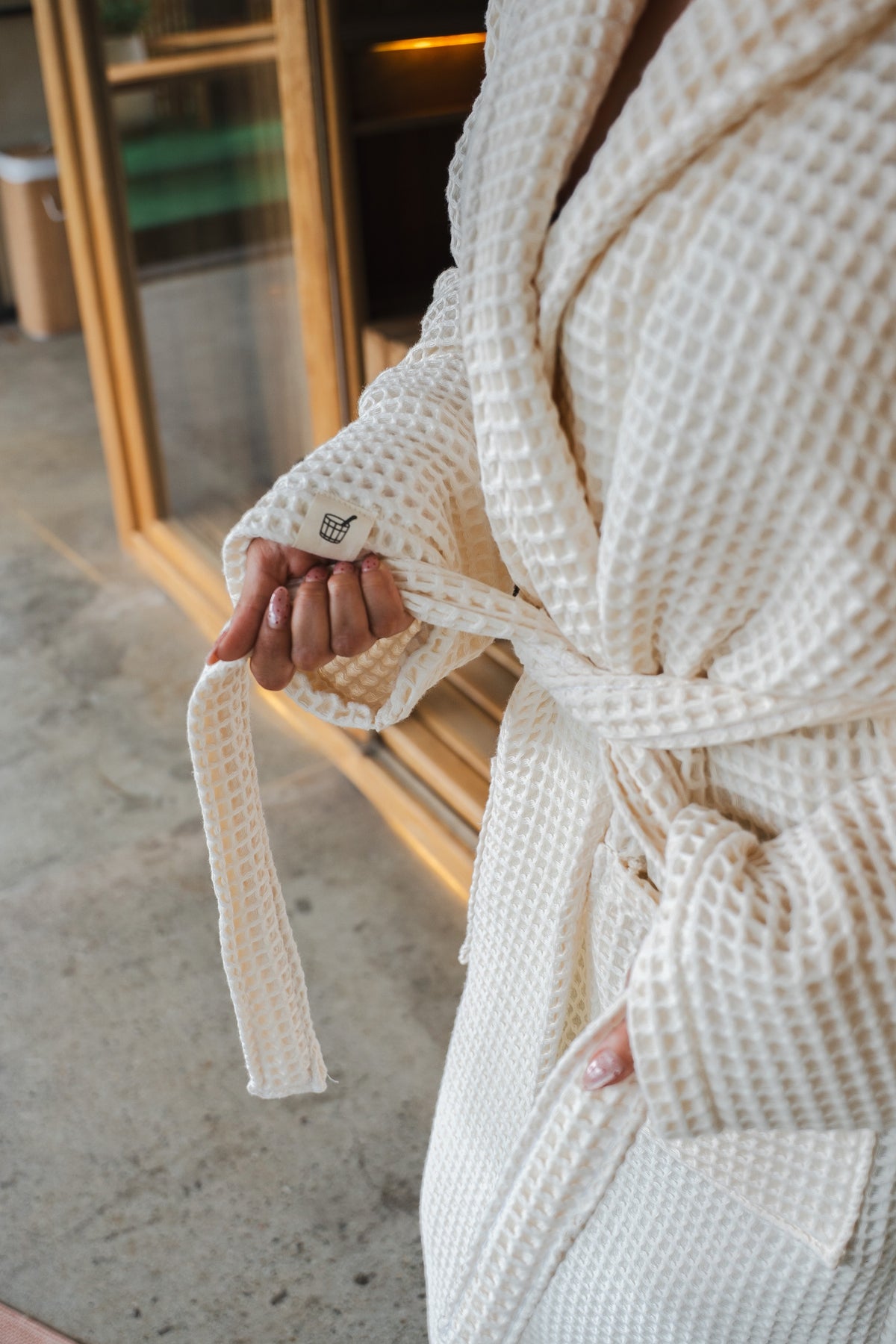 Tying the belt of The Sauna Robe in Arctic Ivory, waffle weave with woven Cult of Sauna cuff label