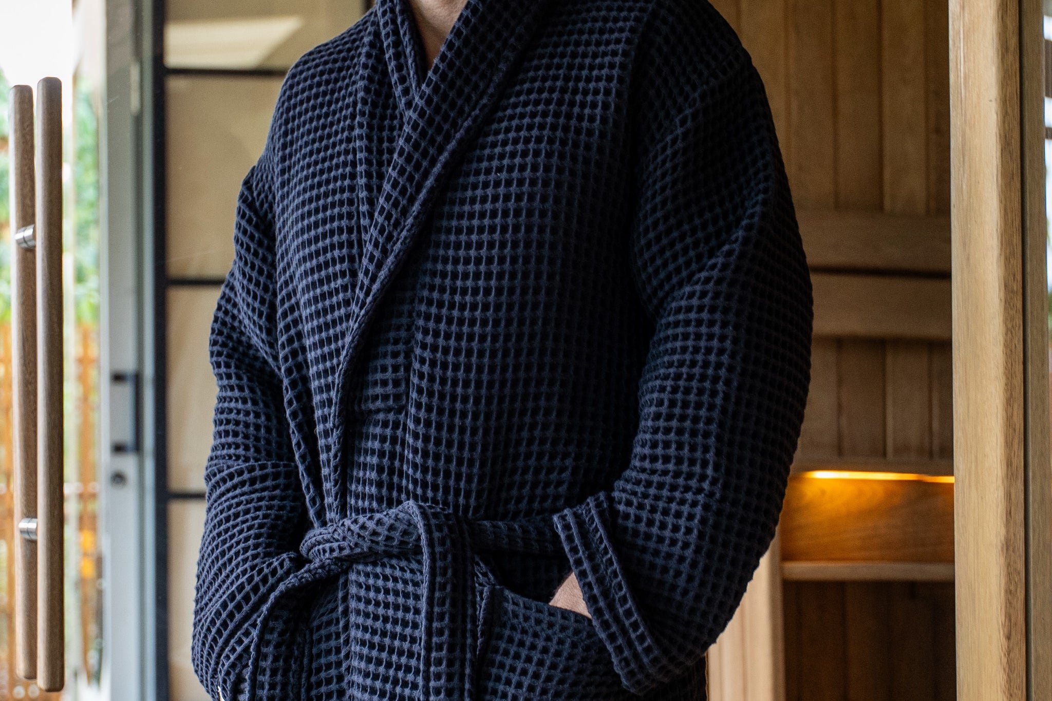 The Sauna Robe in Aurora Black, waffle cotton with shawl collar, worn at the sauna door