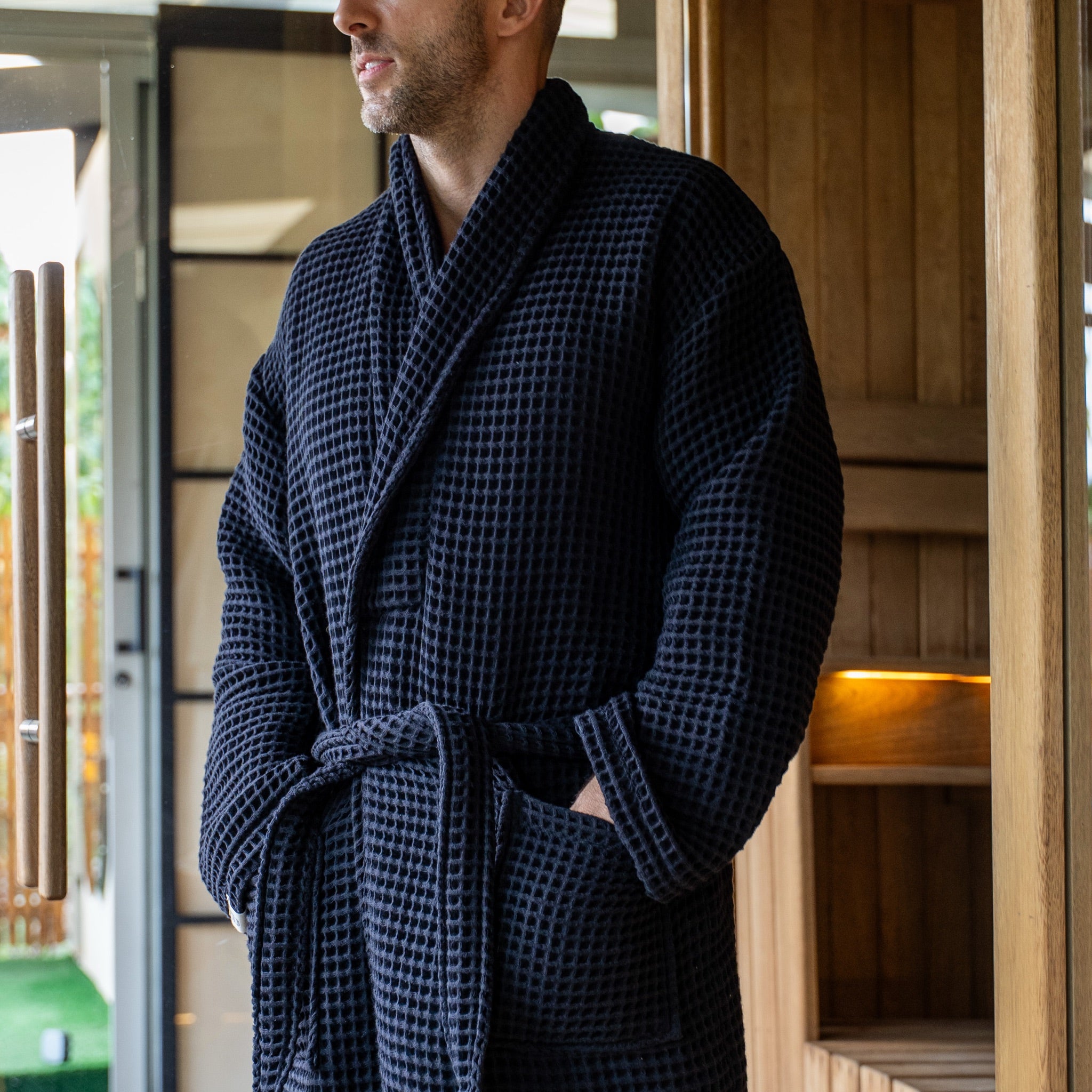 The Sauna Robe in Aurora Black, waffle cotton with shawl collar, worn at the sauna door