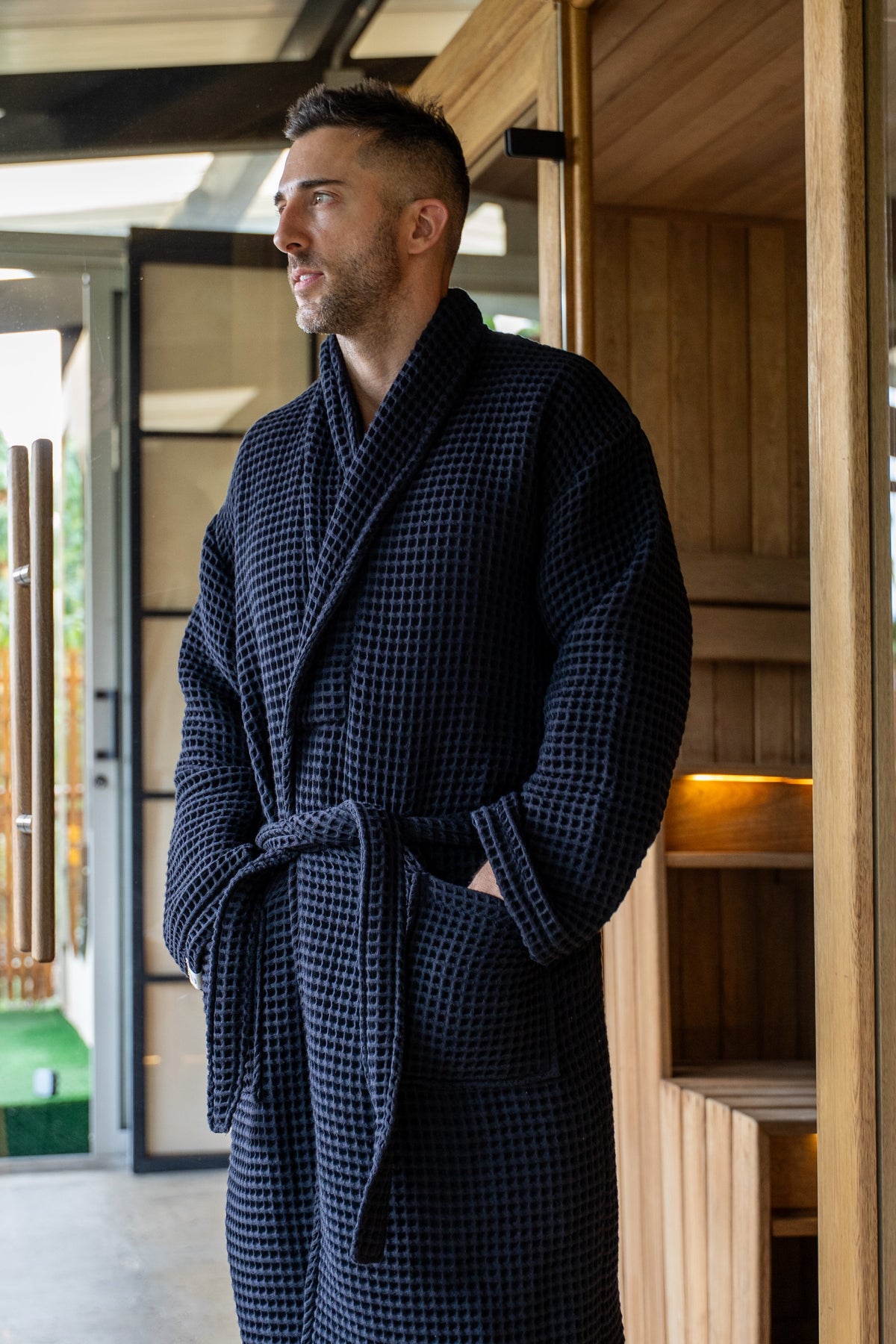 The Sauna Robe in Aurora Black, waffle cotton with shawl collar, worn at the sauna door